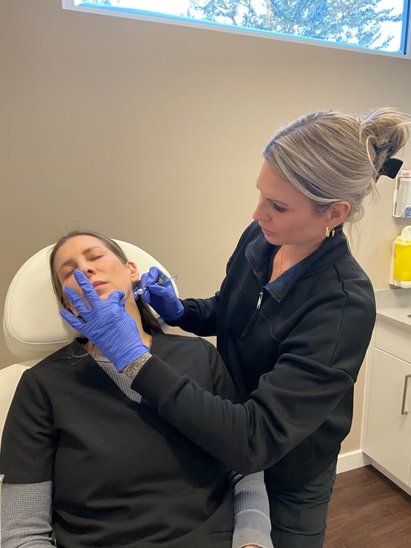 a-medical-professional-in-black-uniform-administering-dermal-fillers-saskatoon-treatment-to-a-patient-sitting-in-a-clinic-chair-wearing-blue-gloves