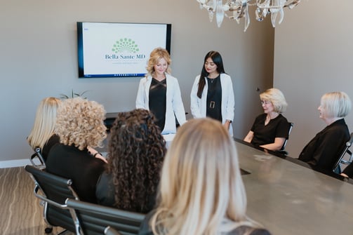 bella-sante-md-staff-meeting-in-conference-room-with-two-women-in-white-coats-standing-and-team-members-seated-around-table-facing-presentation-screen