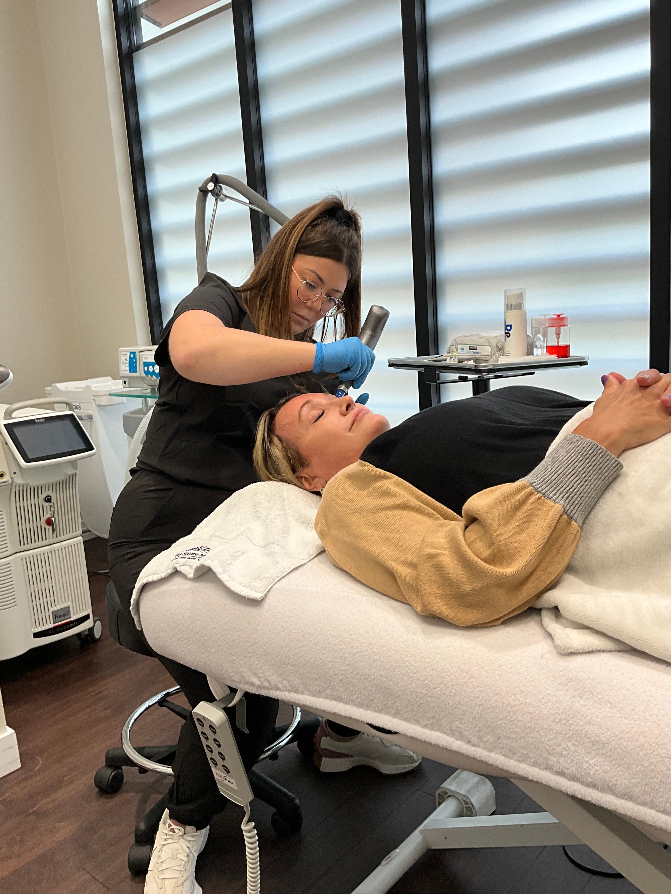 esthetician-performing-skin-treatment-on-client-lying-on-treatment-bed-in-modern-clinic-with-medical-equipment-and-natural-lighting