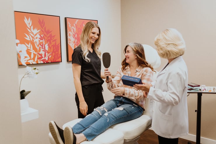 photo-of-woman-in-clinic-chair-holding-mirror-during-co2-laser-before-and-after-consultation-with-two-providers-in-treatment-room