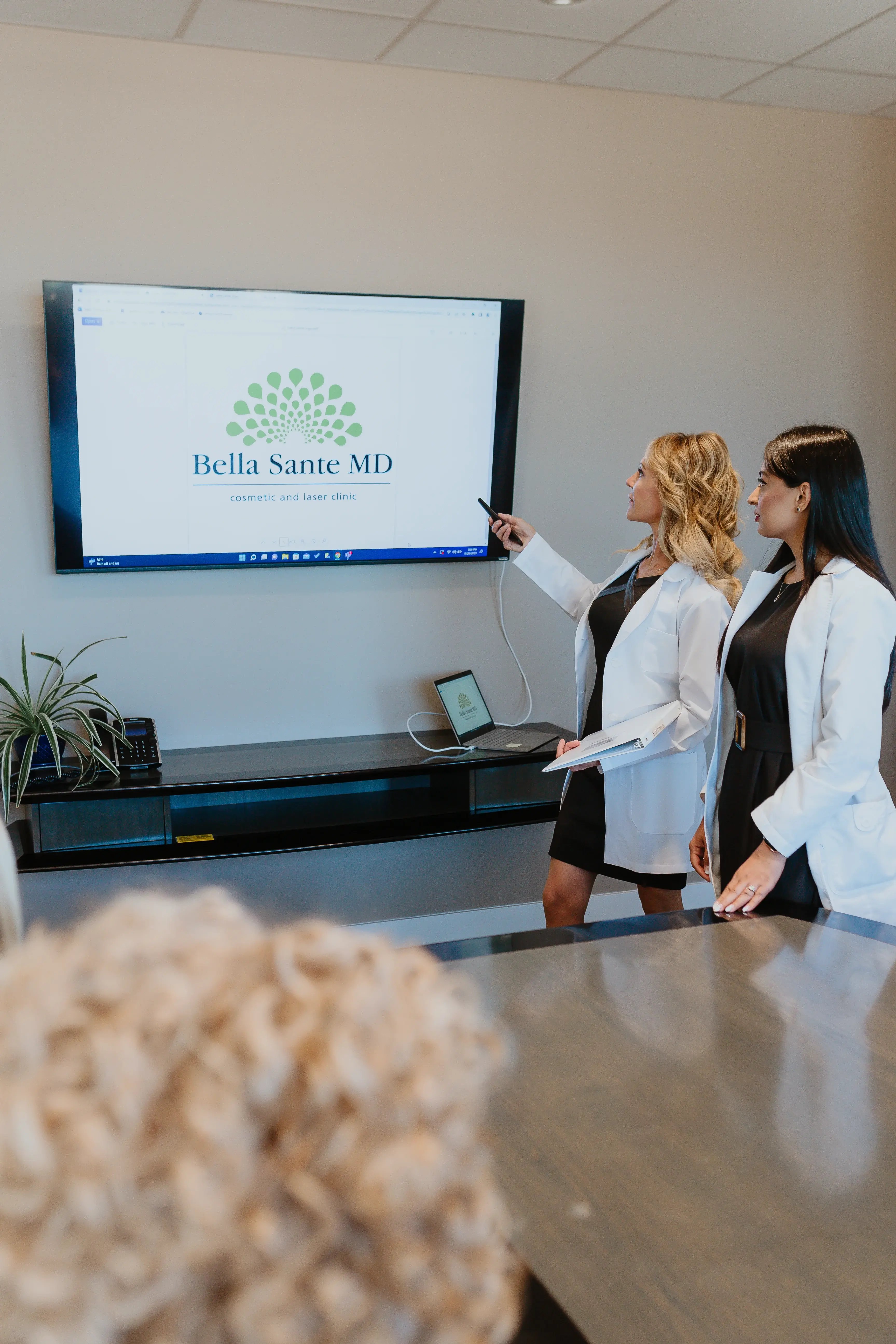 two-women-in-lab-coats-standing-in-office-pointing-at-tv-screen-displaying-bella-sante-md-logo-during-meeting