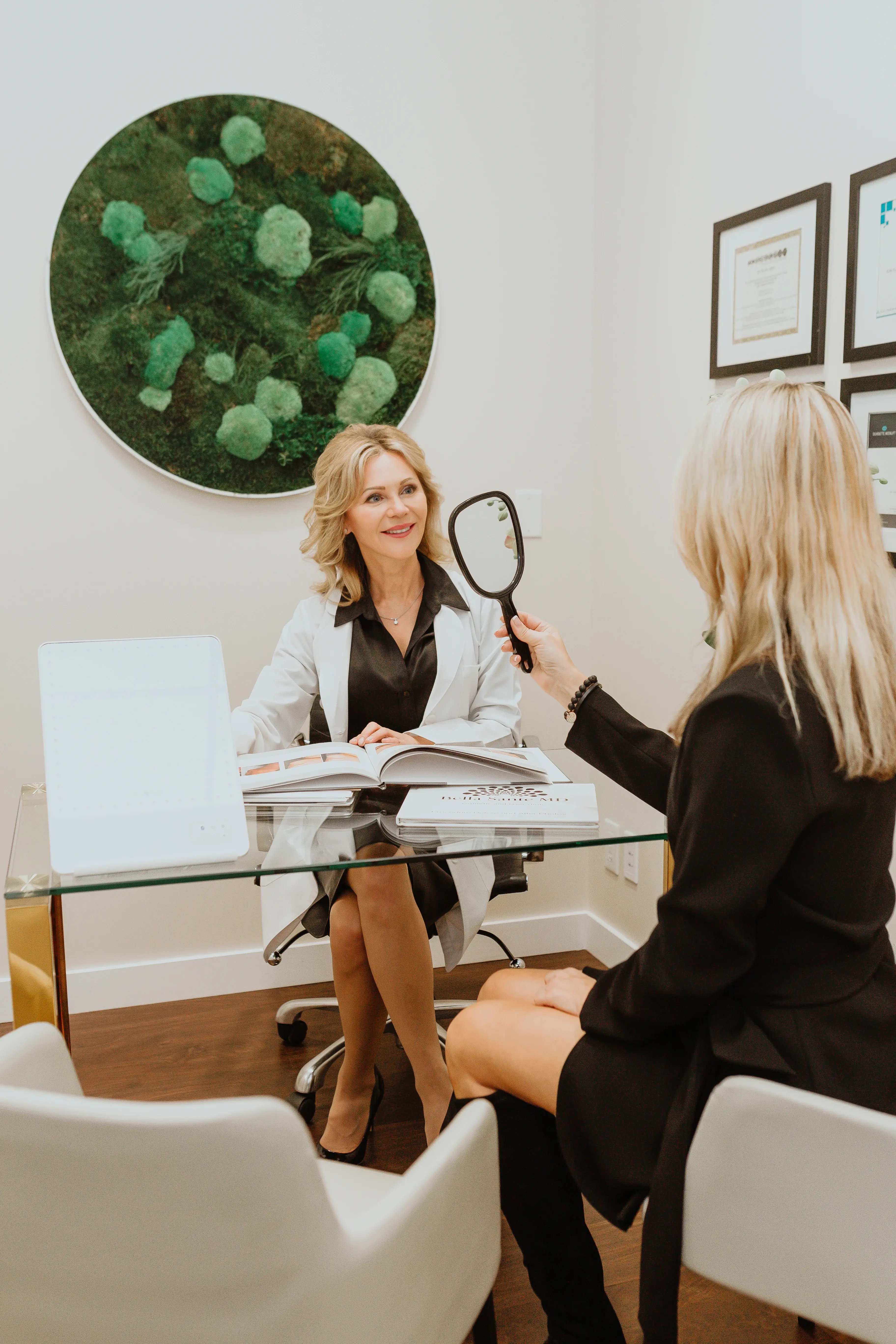woman-in-white-coat-consulting-with-client-holding-hand-mirror-at-desk-in-modern-clinic-setting-thermage-saskatoon-consultation