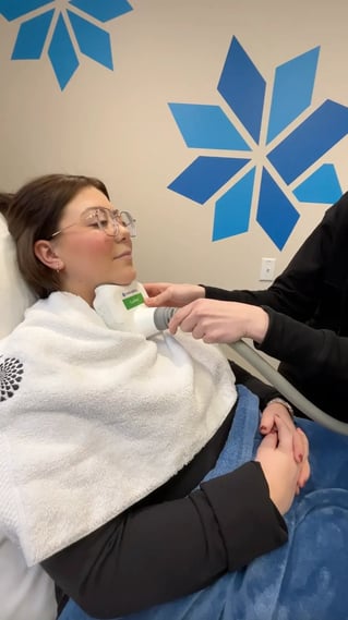 woman-receiving-neck-treatment-with-device-in-clinic-body-contouring-saskatoon-session-blue-wall-designs-in-background-white-towel-and-blanket-for-comfort