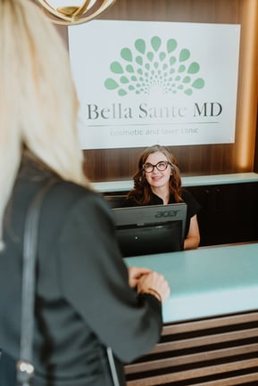 woman-standing-at-reception-desk-speaking-to-smiling-receptionist-with-bella-sante-md-sign-in-background-inside-cosmetic-and-laser-clinic